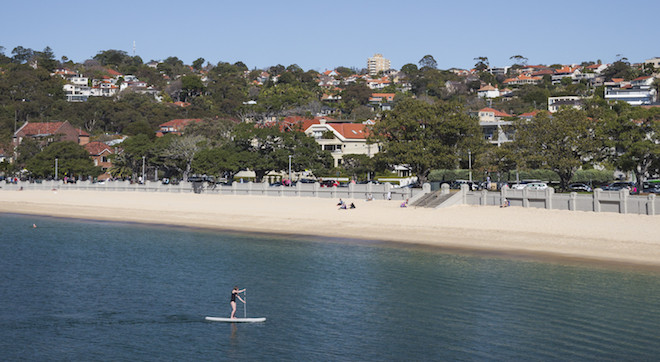 A Stand Up Paddle-boarder paddles back to Balmoral Beach, Mosman. A couple of people are on the beach and some walkers on the Manly to Bondi walk watch from the track behind the beach.