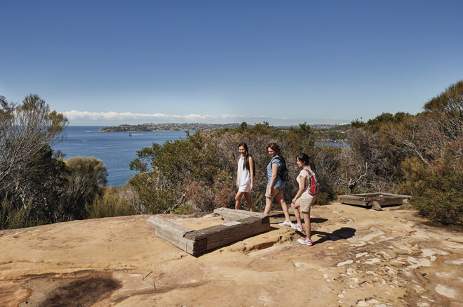 A group of friends admiring the Aboriginal engravings at Grotto Point on the Manly to Spit Bridge walk. Sydney Harbour is visible through the trees in the background.