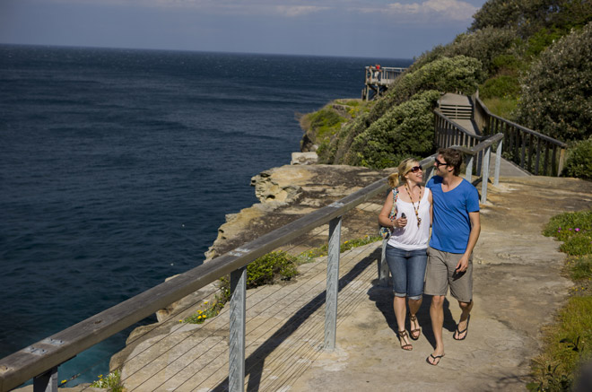 A young couple enjoying a coastal walk along the Federation Cliff Walk from Watsons Bay to Bondi, Sydney. They are walking along the cliff edge near Dover Heights, behind a fence with the ocean behind them on a clear Sydney day.