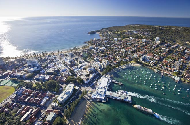 An aerial shot of Manly and North Head. A ferry is approaching the harbour wharf in the bottom of the picture and the ocean stretches to the horizon on the other side of the peninsular.