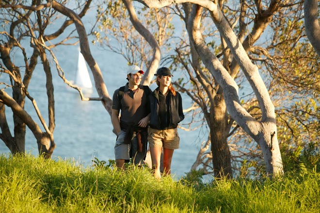 A couple walking through trees on the Bondi to Manly walk. A boat sailing on the harbour is visible through the trees behind them as they enjoy one of the best walks in Sydney on a late, sunny afternoon.